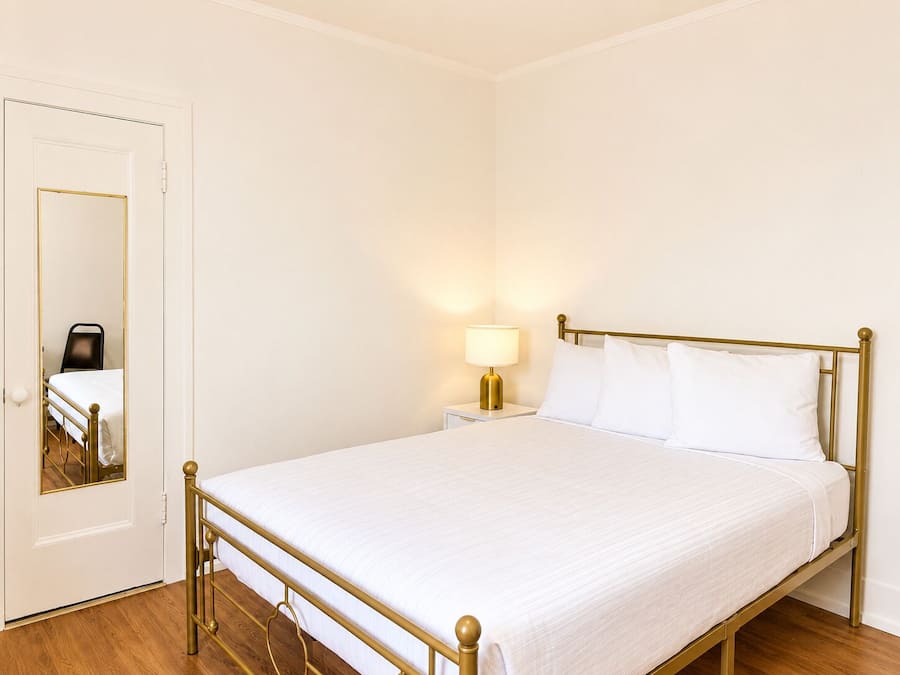 Quarto individual com uma cama, piso de madeira, mesa de cabeceira com abajur e porta com espelho.