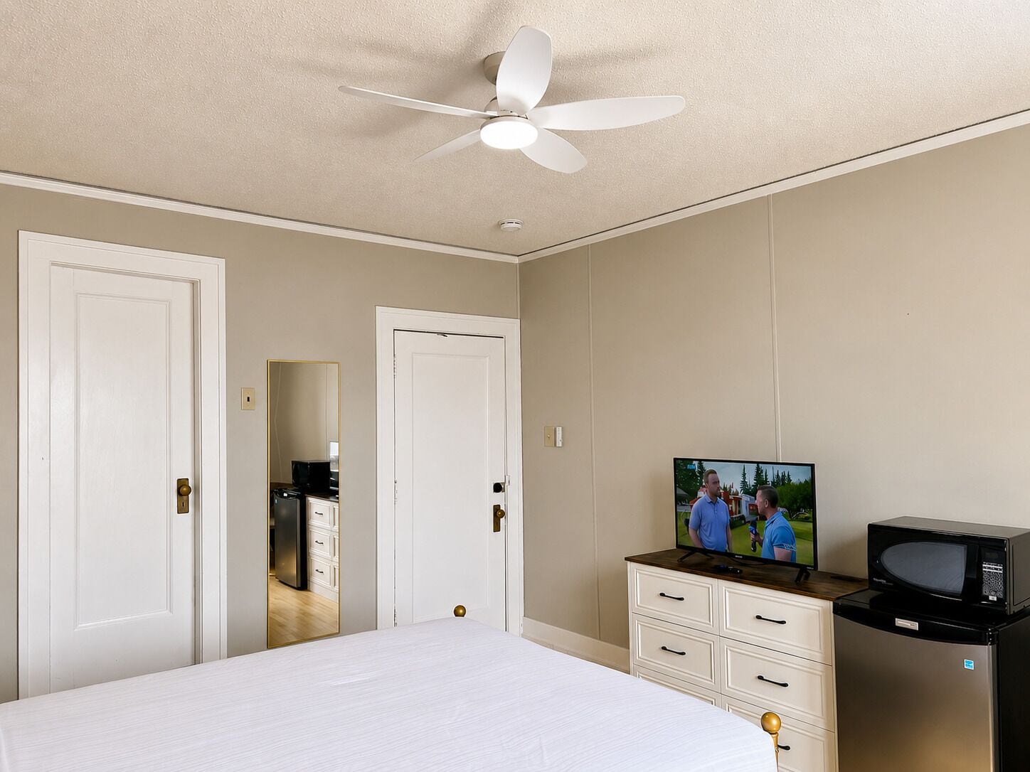 A bedroom with a bed, a television, a microwave, and a ceiling fan.