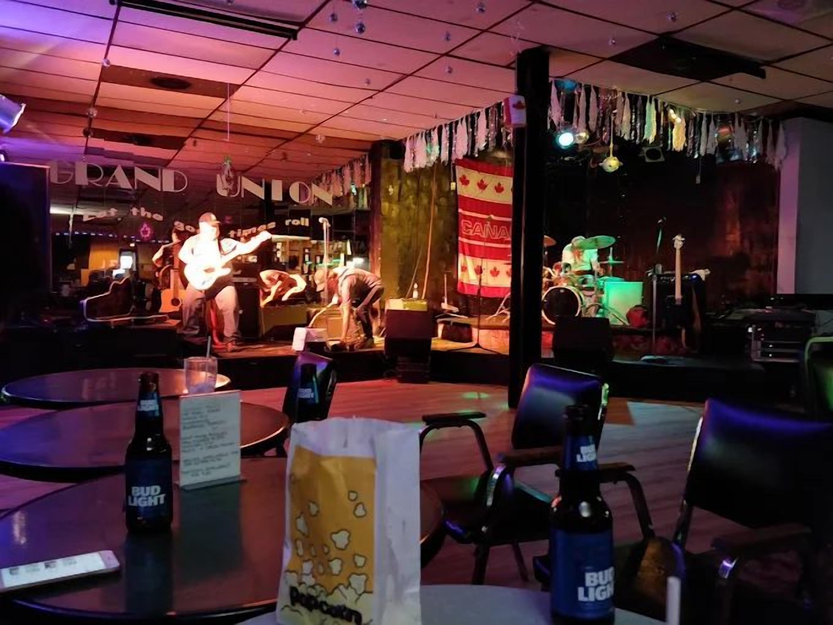 Uma área de bar com palco para música ao vivo, mesas com cadeiras e uma garrafa de Bud Light sobre uma mesa.