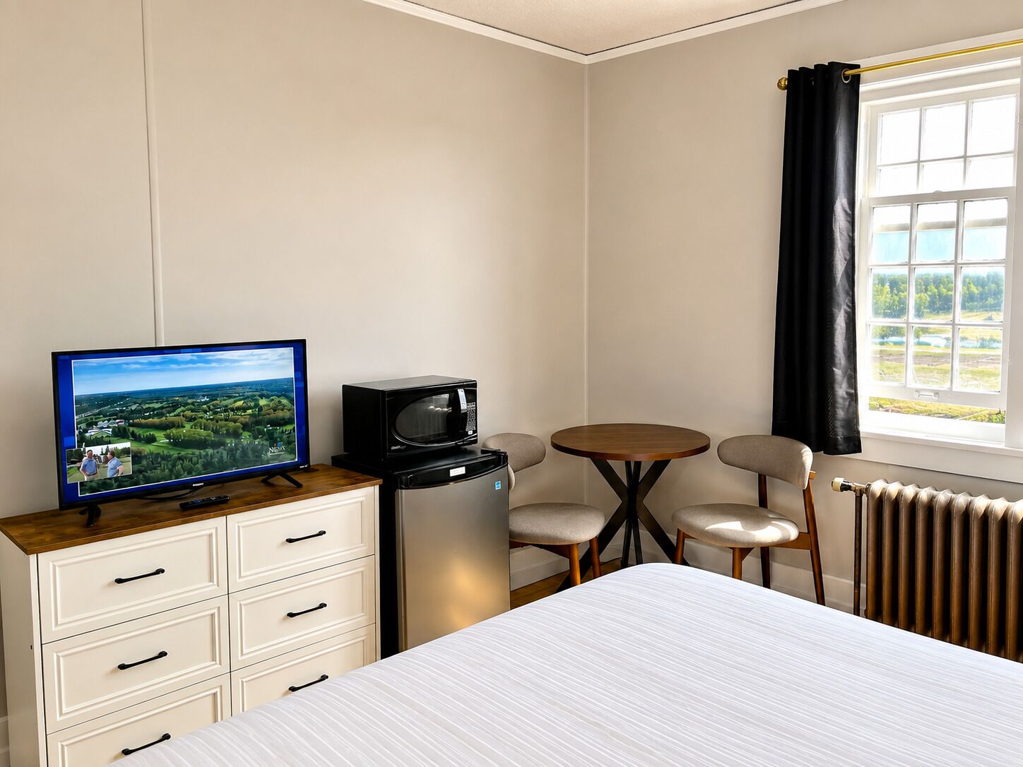 A hotel room with a bed, a television, a small table with chairs, a dresser, and a window with curtains.