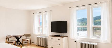 A hotel room with a bed, a desk with a chair, a television, and two windows with curtains.