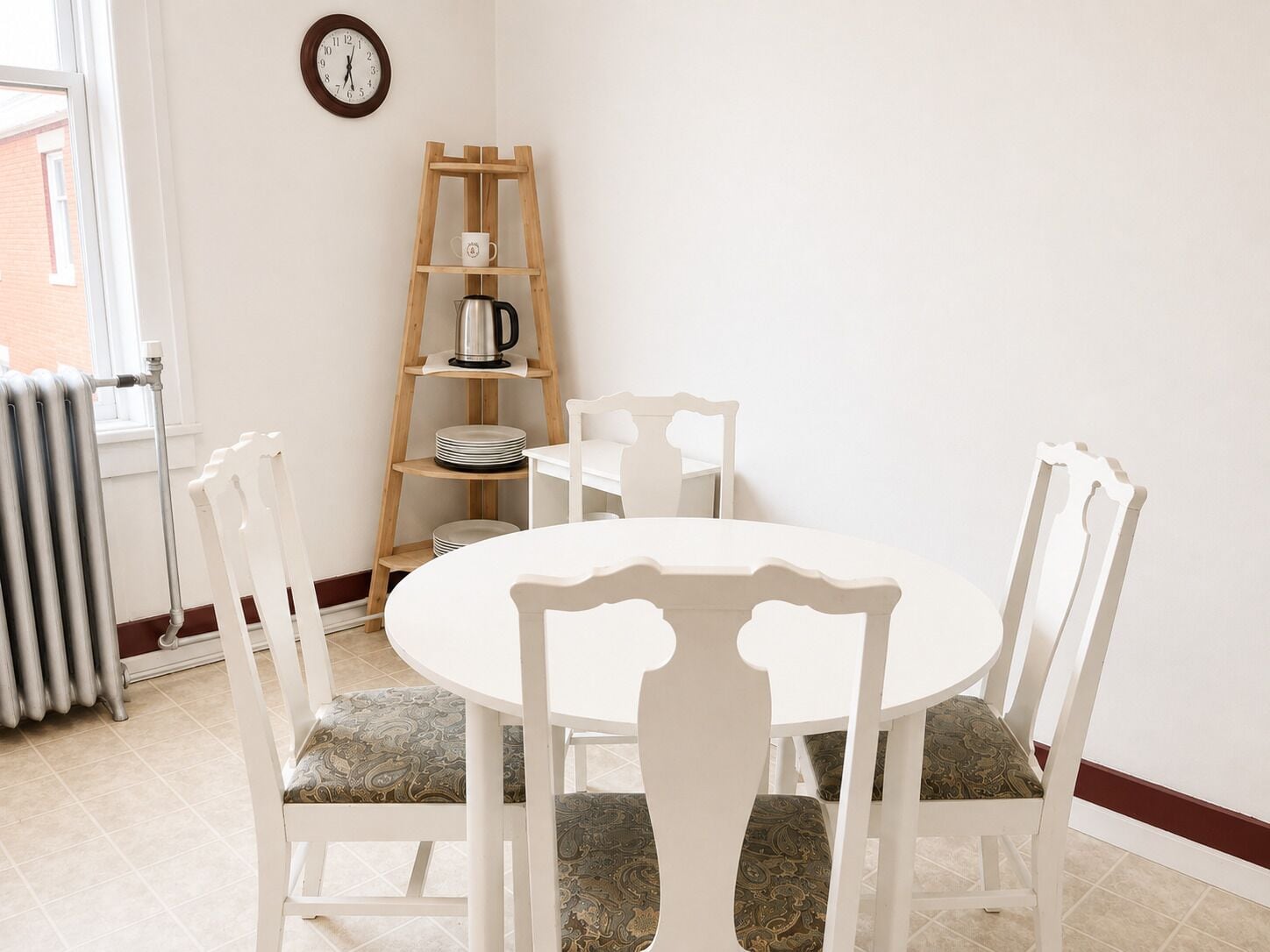 Un coin repas avec une table ronde, des chaises blanches, une étagère en bois sur laquelle se trouve une bouilloire, et une horloge au mur.