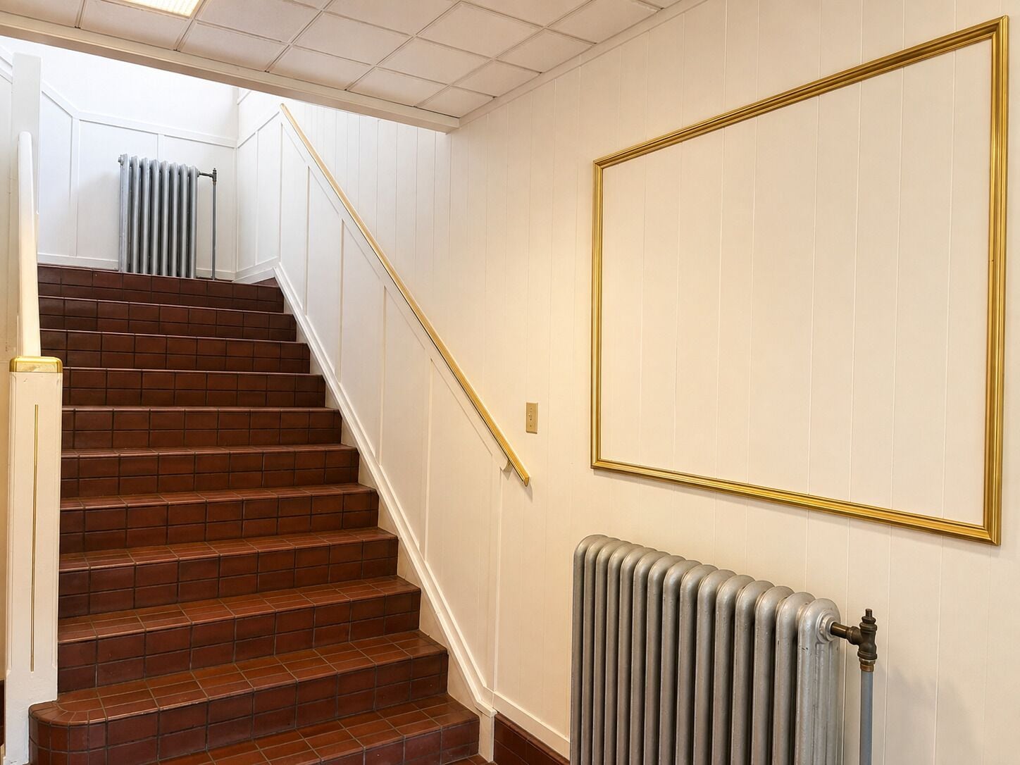 A staircase with brown tiles leading to a landing, a radiator on the landing, and a framed picture on the wall.