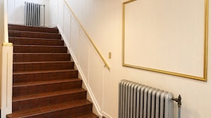 Una escalera con baldosas marrones que lleva a un rellano, un radiador en el rellano y un cuadro en la pared.
