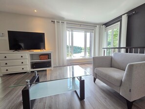 A modern living room with a flat-screen TV, a glass coffee table, a grey sofa, and a large window with curtains.