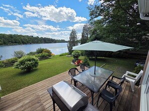 A deck with a grill, chairs, and a table with an umbrella, overlooking a lake with boats.