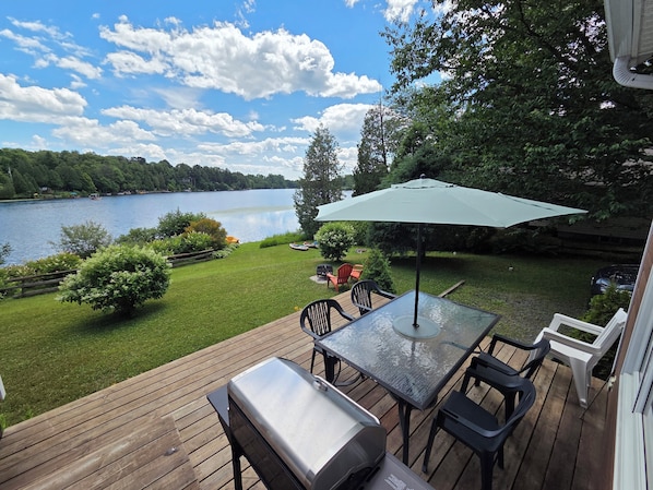 A deck with a grill, chairs, and a table with an umbrella, overlooking a lake with boats.