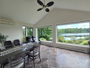 A room with a stone floor, a table with chairs, a ceiling fan, and large windows overlooking a lake.