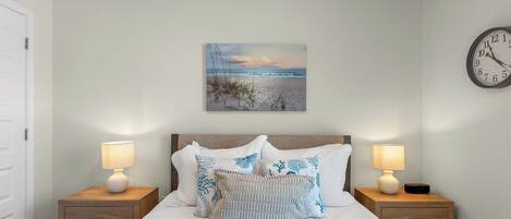 A bedroom with a bed, two bedside tables, a wall clock, and a beach-themed painting.