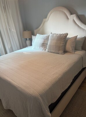 Uma cama com cabeceira branca e várias almofadas, abajur e janela com cortinas.