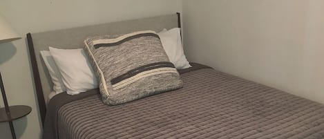 A bed with a grey quilt, two pillows, a bedside lamp, and a patterned rug.