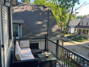 A balcony with wicker furniture and a glass table, overlooking a street with houses and trees. - Vintage Art Lodge 4BR/2BA Full Home in Bay View (Milwaukee)