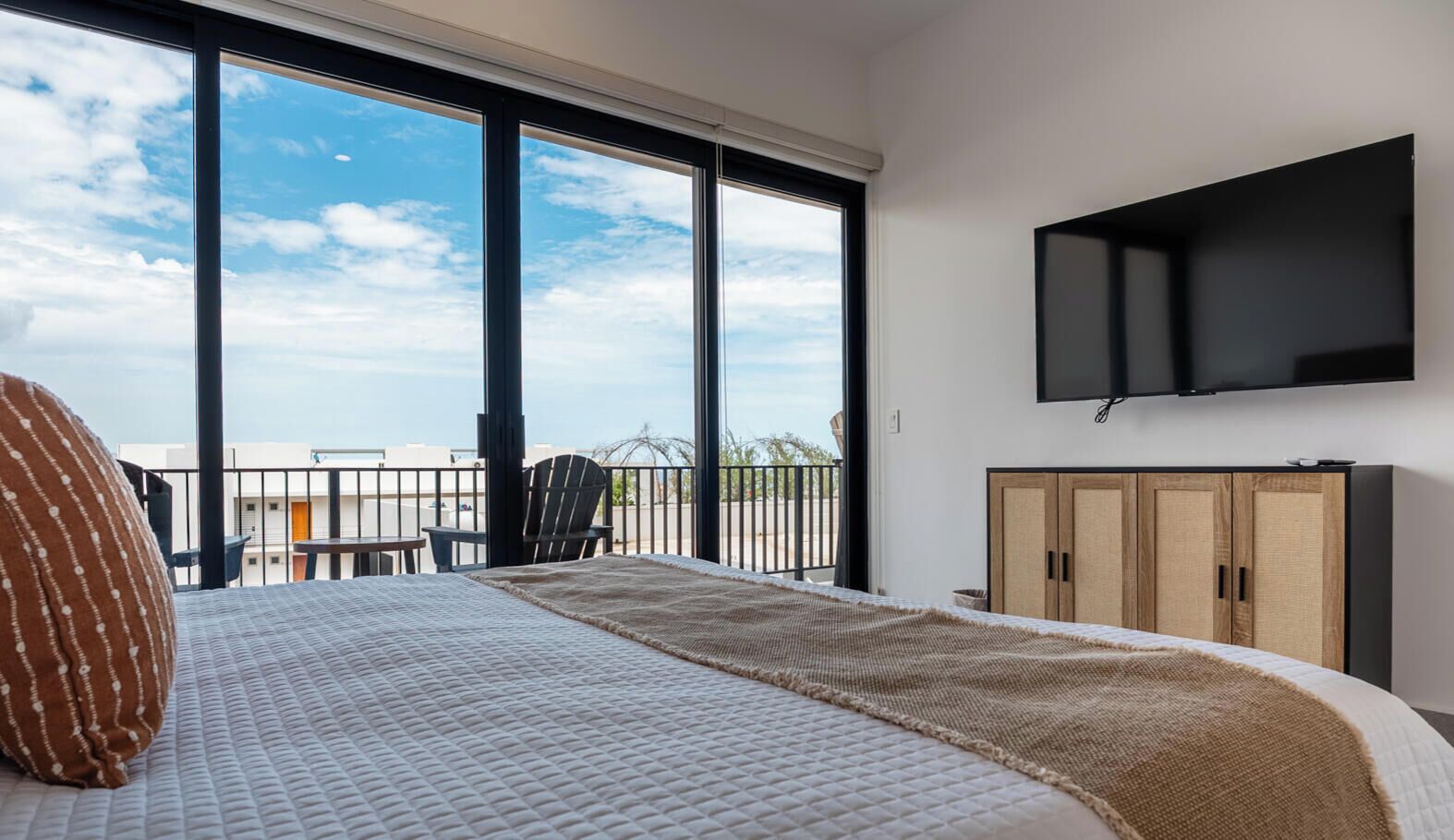 Un dormitorio con cama, televisor montado en la pared y vistas al exterior a través de amplias ventanas.