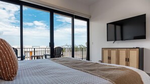 A bedroom with a bed, a television mounted on the wall, and a view of the outside through large windows. - Palma Blanca 2BR Condos with Rooftop Pool and Ocean Views by Baja Sol PM (Cabo San Lucas)