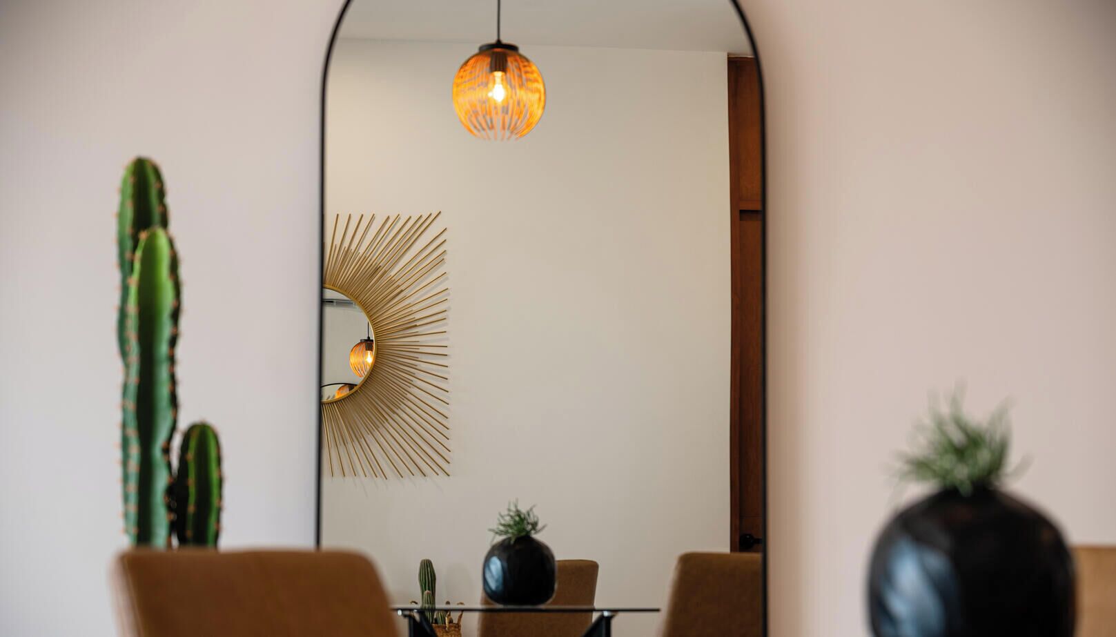 A modern interior with a dining area, a cactus plant, a round mirror, and a hanging light fixture.