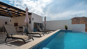 Una piscina al aire libre con sillones de descanso y sombrillas, una zona de bar y vistas a una colina.