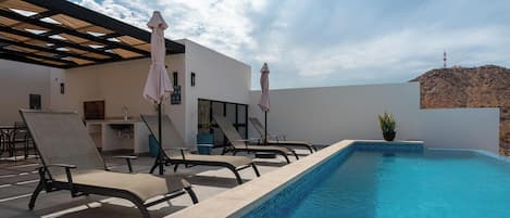 Una piscina al aire libre con sillones de descanso y sombrillas, una zona de bar y vistas a una colina.