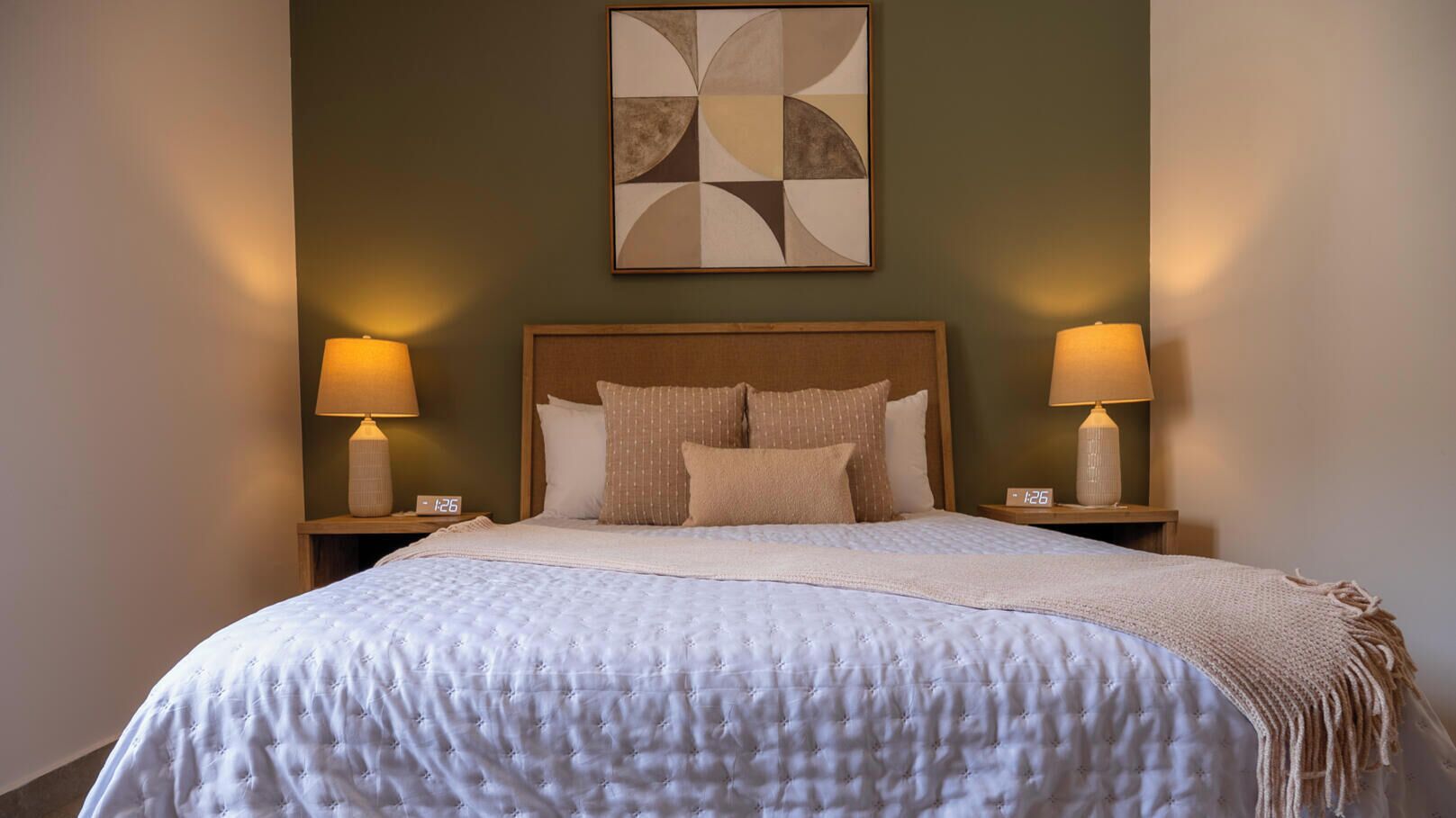 A neatly made bed with a white duvet and pillows, two bedside lamps, a clock, and a geometric abstract painting on the wall.