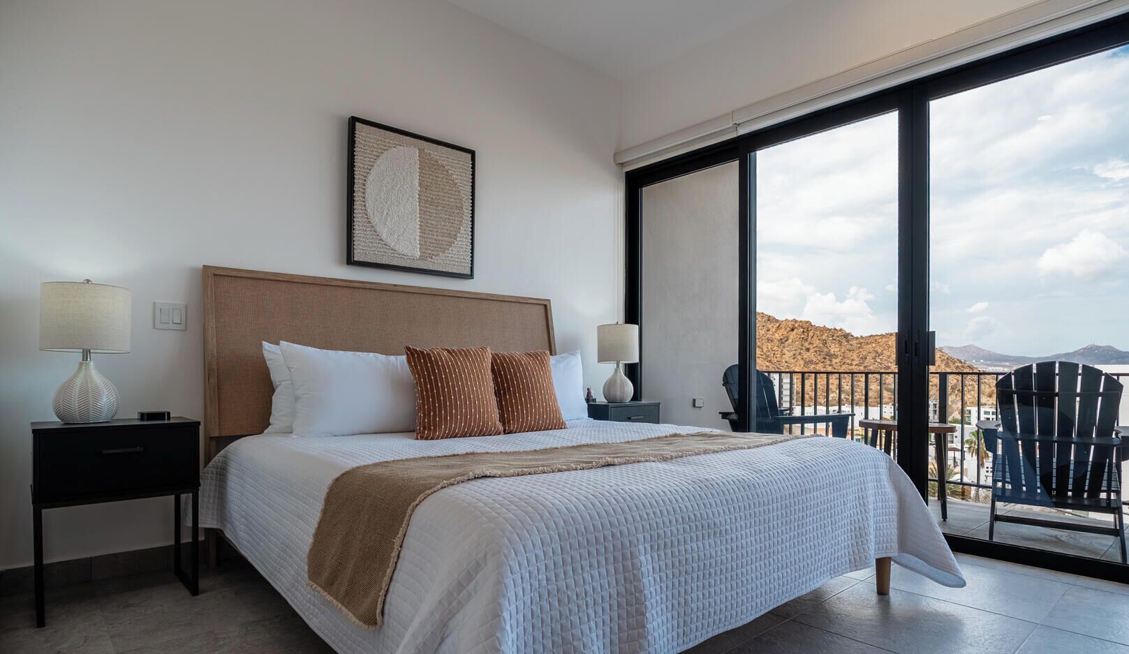 A hotel room with a bed, two bedside tables with lamps, a large window with a view of the landscape, and a balcony with chairs.