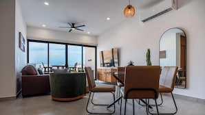 A modern living room with a sofa, chairs, a dining table, and a large window offering a view. - Palma Blanca 2BR Condos with Rooftop Pool and Ocean Views by Baja Sol PM (Cabo San Lucas)