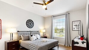 A bedroom with a bed, bedside tables, a ceiling fan, a window with curtains, and a wall decoration. - Coconico Retreat At Windsor Palms (Kissimmee)