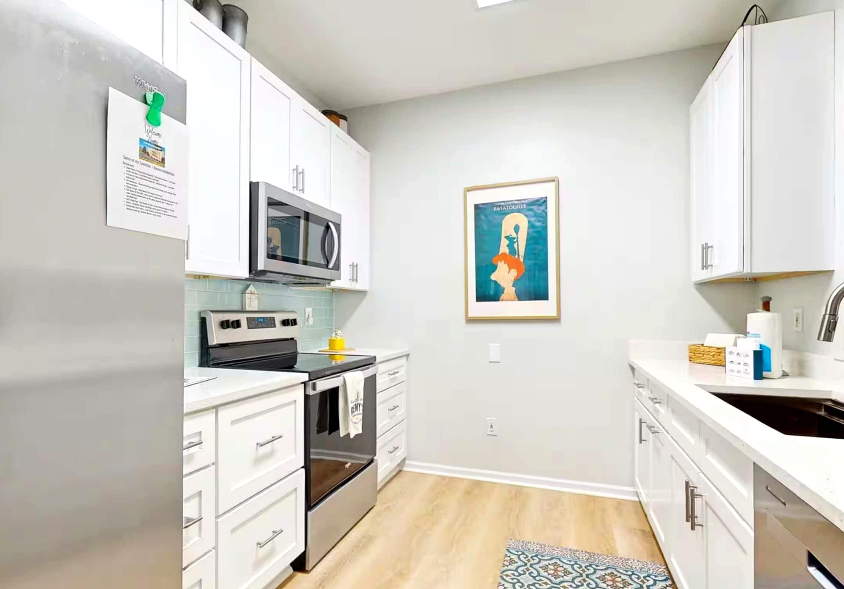 A modern kitchen with white cabinets, a microwave, an oven, and a sink. There is a framed picture on the wall and a small rug on the floor.