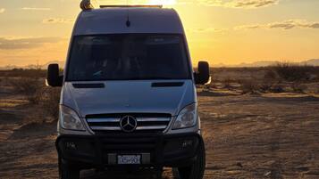 Un fourgon Mercedes-Benz blanc est garé sur un chemin de terre, avec un coucher de soleil en arrière-plan.