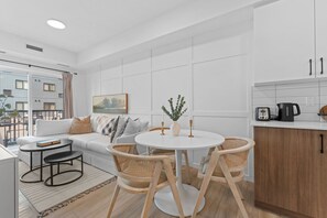 Ein modernes Wohnzimmer mit einer Couch, einem runden Esstisch und Stühlen. Es gibt eine Küchenzeile mit Schränken und einem Wasserkocher.