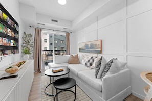 A modern living room with a white sofa, a small round table, and a bookshelf filled with DVDs. - The Willow | Boutique 1BR w/ Balcony (Edmonton)