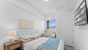 Un dormitorio con cama, mesitas de noche, lámpara, televisor montado en la pared y ventana con vistas.