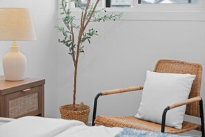 Ein Korbstuhl mit weißem Kissen, ein Nachttisch mit Lampe und ein Topfpflanze.