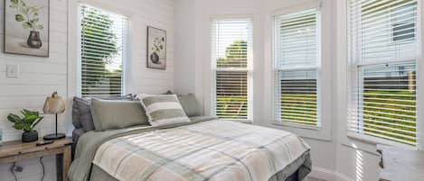 A bedroom with a bed, a desk, and a chair. There are three windows with blinds.