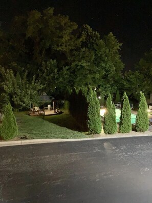 Vista nocturna de un jardín con piscina, rodeado de árboles y un patio con mobiliario exterior.