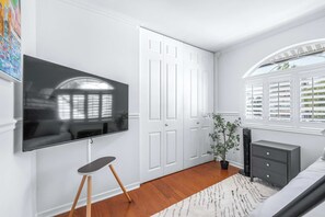 Ein Schlafzimmer mit Flachbildfernseher, einem Fenster mit Jalousien, einem Blumentopf, einer Kommode und einem Teppich.