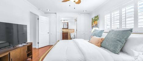 Un dormitorio con cama, televisión, ventilador de techo y baño visible a través de una puerta abierta.