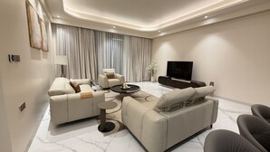 Een moderne woonkamer met een zithoek, een salontafel, een flatscreen televisie en een groot raam met gordijnen.