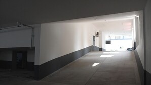 Um corredor com chão de concreto, paredes brancas e rodapé preto. Há uma porta no final do corredor que dá para um quarto iluminado com janelas.