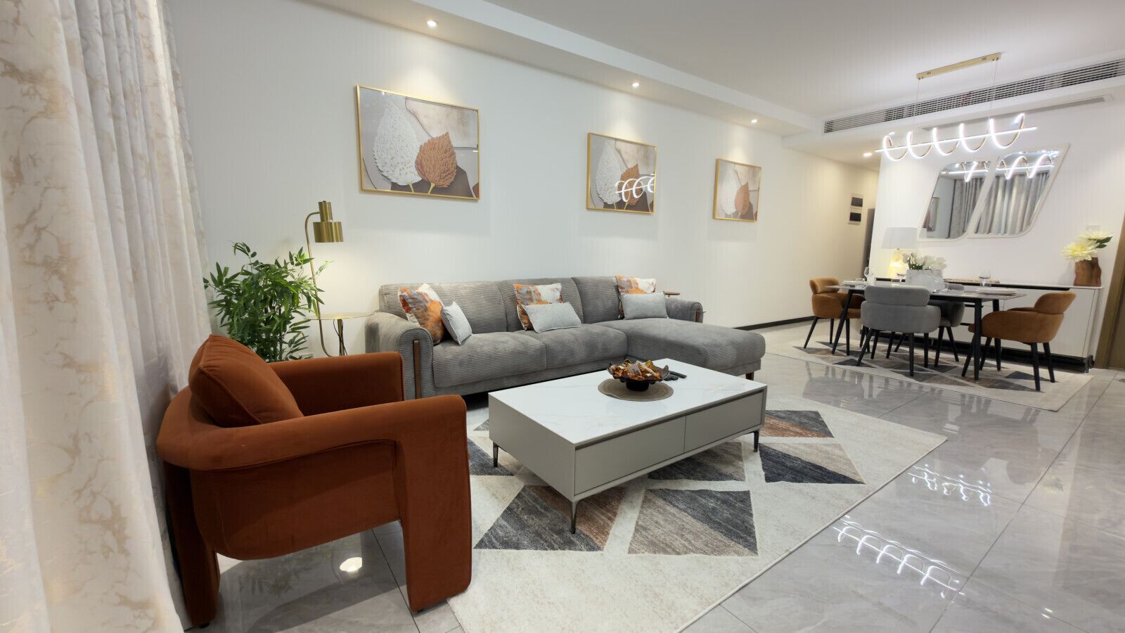 A modern living room with a grey sofa, a brown armchair, and a coffee table. There is a dining area with a table and chairs.