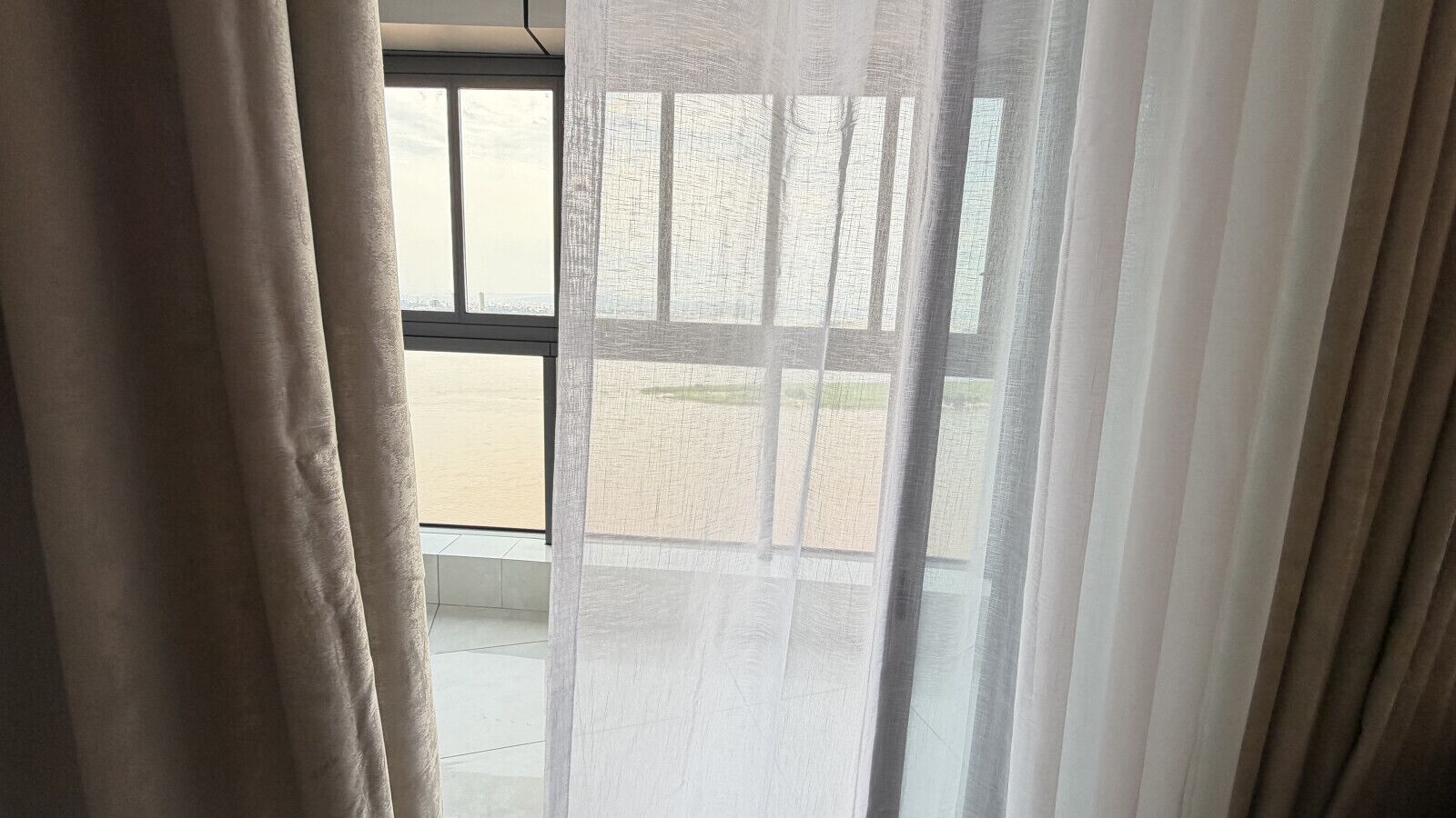 A room with a view through a window, curtains, and a tiled floor.