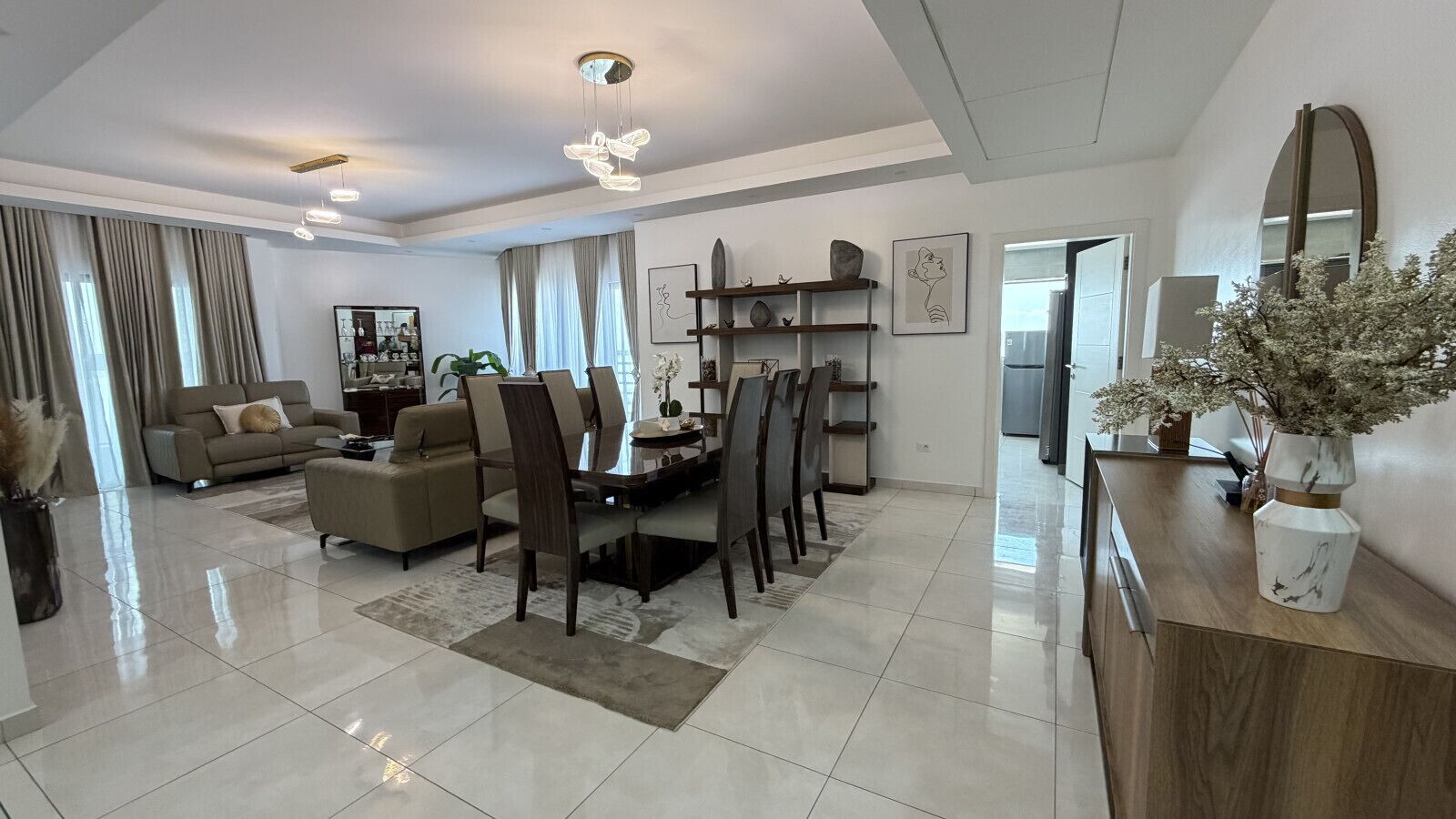 A modern living room with a sofa, dining table, chairs, and a coffee table. There is a kitchen area with cabinets and a refrigerator.