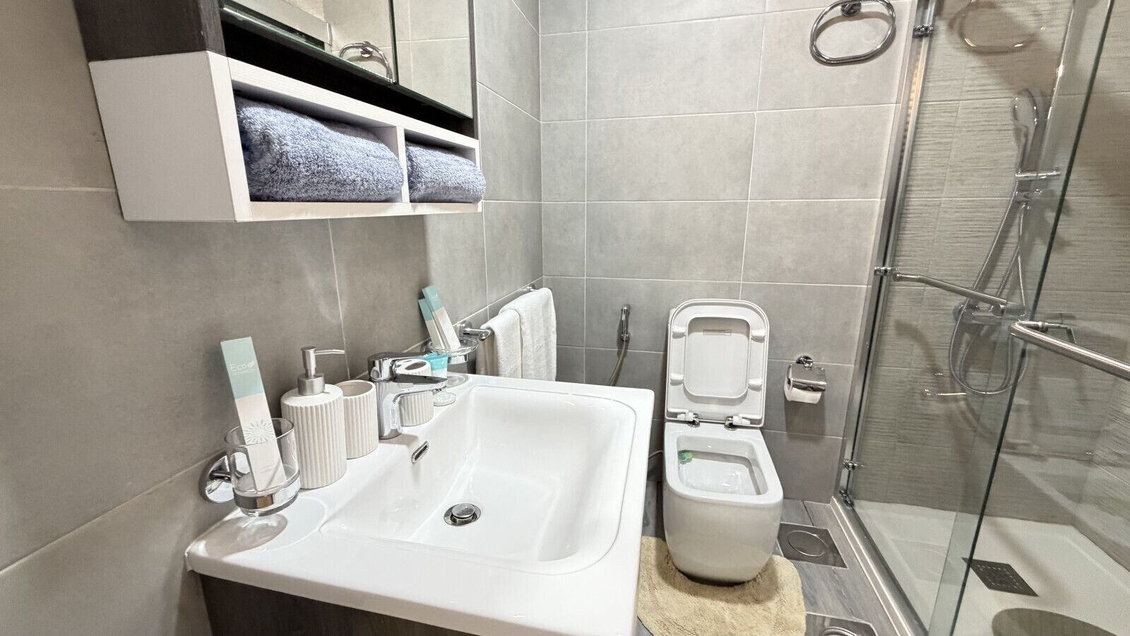 Un baño moderno con un lavamanos blanco, una cabina de ducha con paredes de vidrio y un inodoro con asiento elevado.