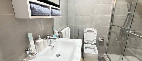 Un baño moderno con un lavamanos blanco, una cabina de ducha con paredes de vidrio y un inodoro con asiento elevado.
