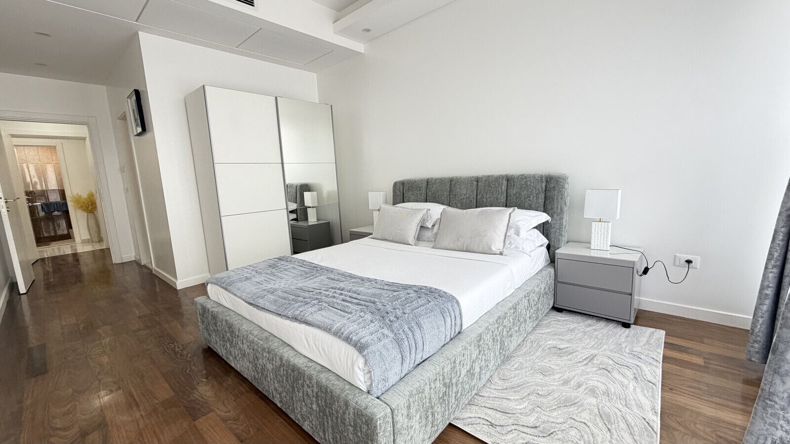 A modern bedroom with a large bed, bedside tables, a wardrobe, and a connecting hallway.
