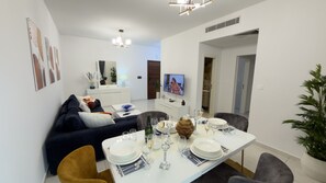 A modern dining area with a white table set for a meal, a sofa, and a television in the background. - Elegant 8th Floor River View Apartment Eloge Tower (Kinshasa)