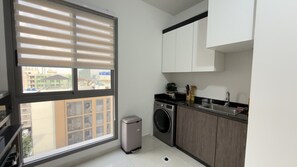 A compact kitchen with a washing machine, a sink, and a window with blinds. - Elegant 8th Floor River View Apartment Eloge Tower (Kinshasa)