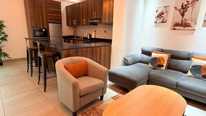 Un saló modern amb un sofà, una butaca i una taula de centre. Es veu una cuina al fons amb armaris de fusta i un frigorífic.