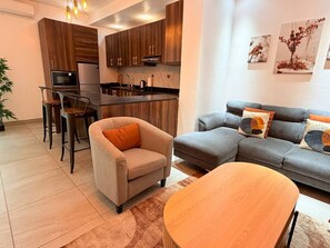A modern living room with a sofa, armchair, and coffee table. A kitchen with wooden cabinets and a refrigerator is visible in the background. - Kintambo Velodrome apartment in a secure building with Gym (Kinshasa)