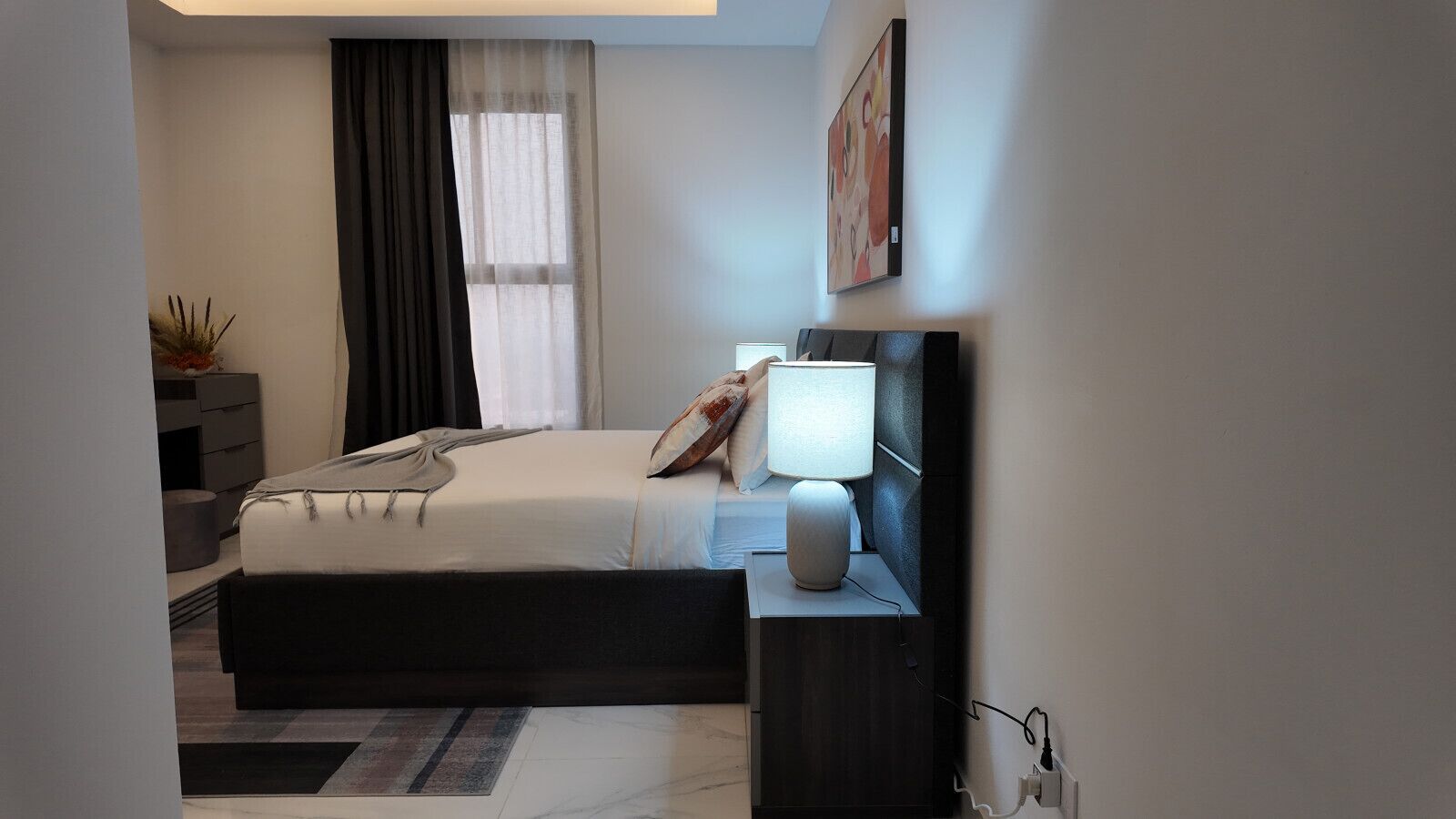 Un dormitorio moderno con cama, mesita de noche, lámpara y una ventana con cortinas.