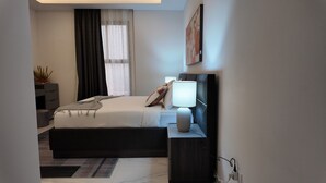 A modern bedroom with a bed, bedside table, lamp, and a window with curtains. - Luxury 3 Bedroom apartment (Kinshasa)