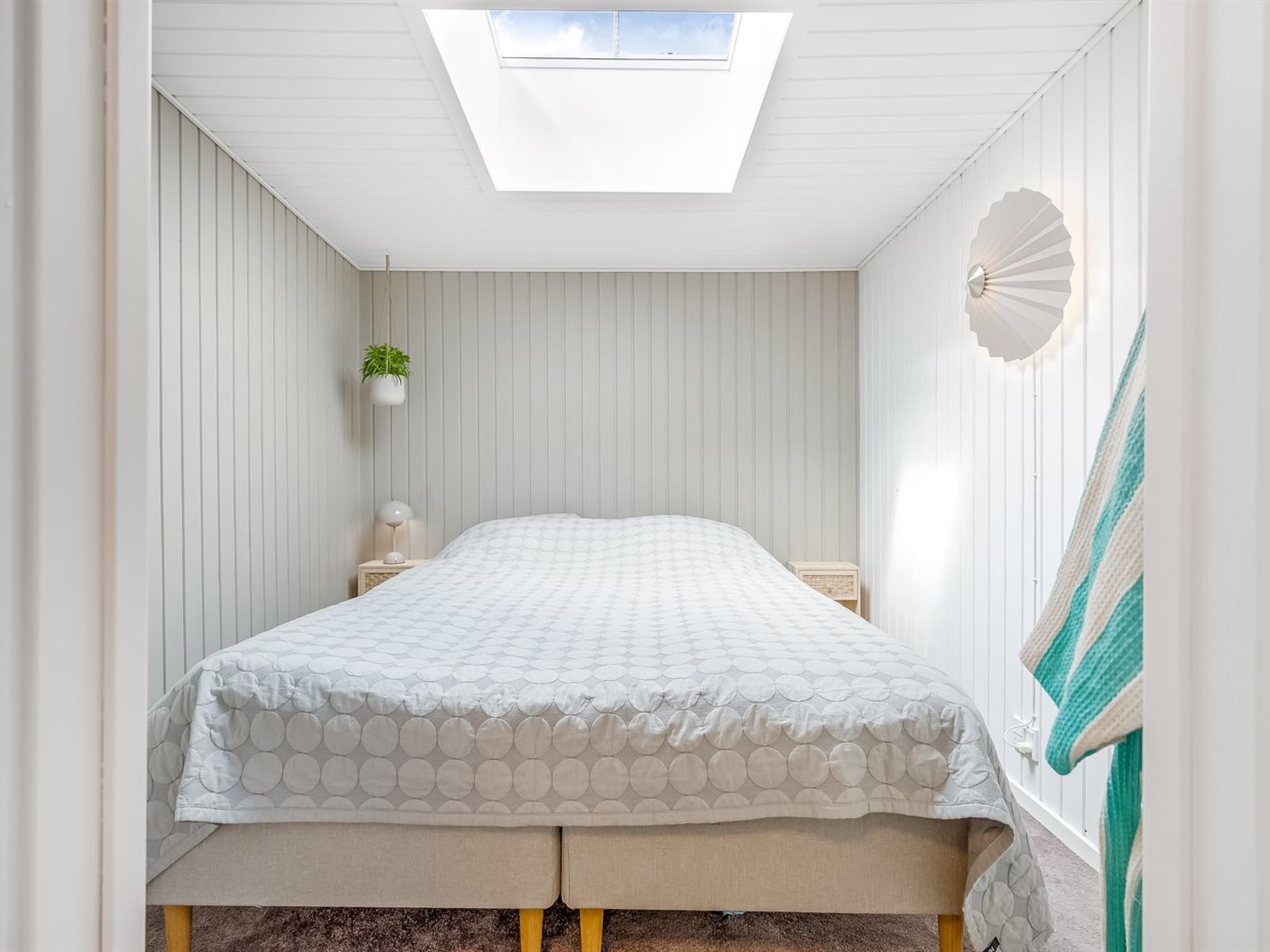 Une chambre avec un puits de lumière, un lit recouvert d’une couvre-lit à motifs circulaires, une table de chevet avec une lampe et un ventilateur fixé au mur.